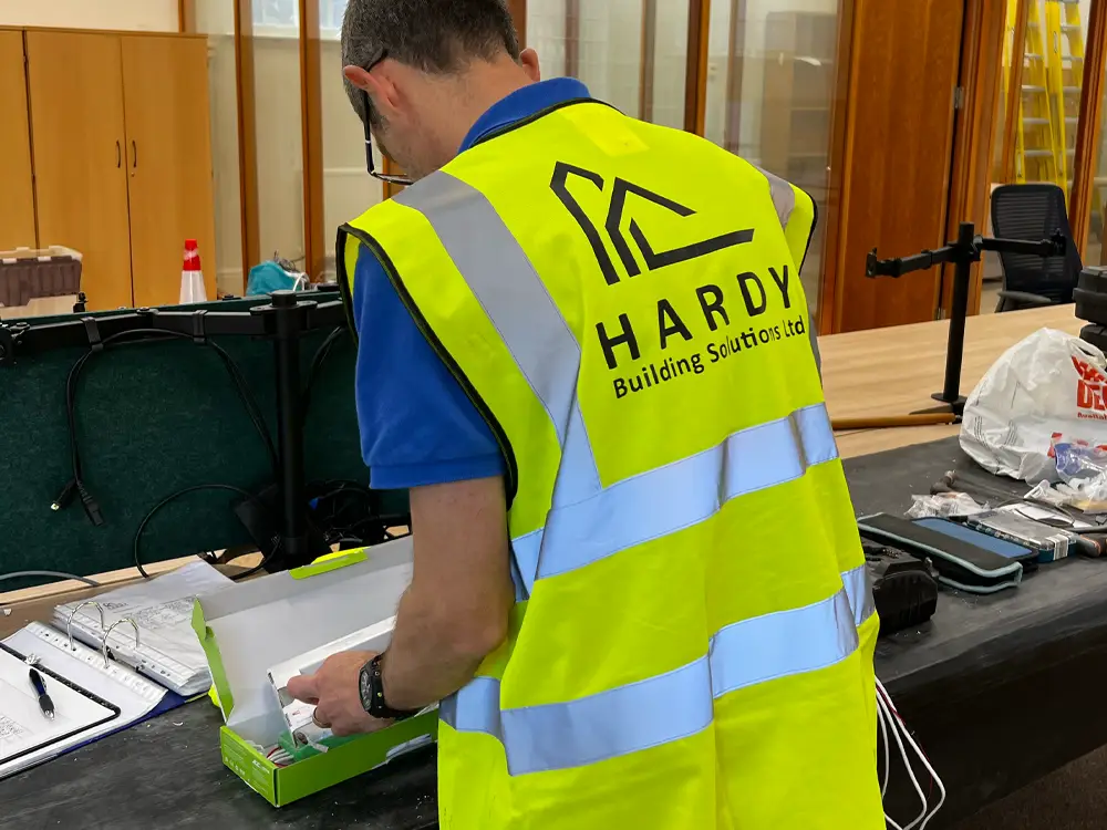 Image shows a man wearing hi vis with Hardy Building Solutions logo on the back standing at a desk unboxing electrical equipment